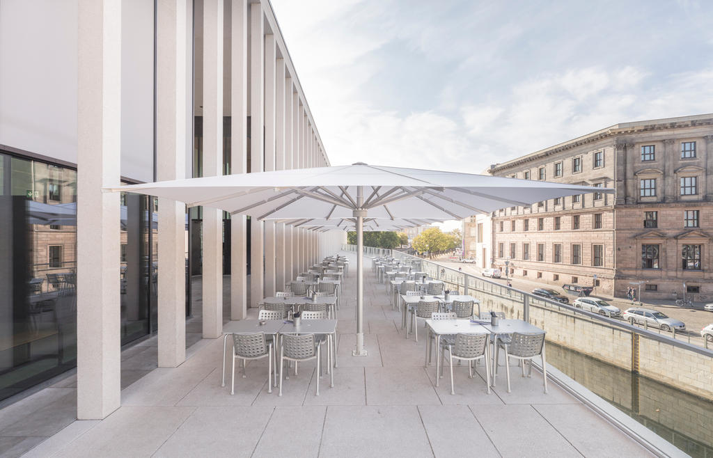 Museumsinsel, James-Simon-Galerie weißen ALBATROS Schirmen Eine moderne Außenterrasse mit weißen Sonnenschirmen, Tischen und Stühlen entlang eines Kanals neben einem Gebäude
