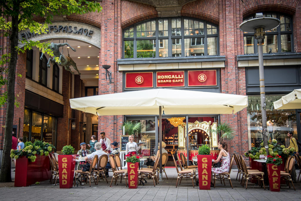 Roncalli Grand Café mit beigen SCHATTELLO Schirmen eine Café-Terrasse mit Sonnenschirmen vor einem historischen Backsteingebäude