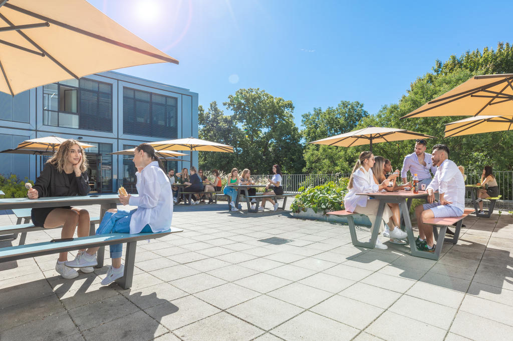 State vocational school, Landshuth with beige SCHATTELLO parasols A terrace with beige parasols where people are sitting, in the background a historic town with a church tower and castle
