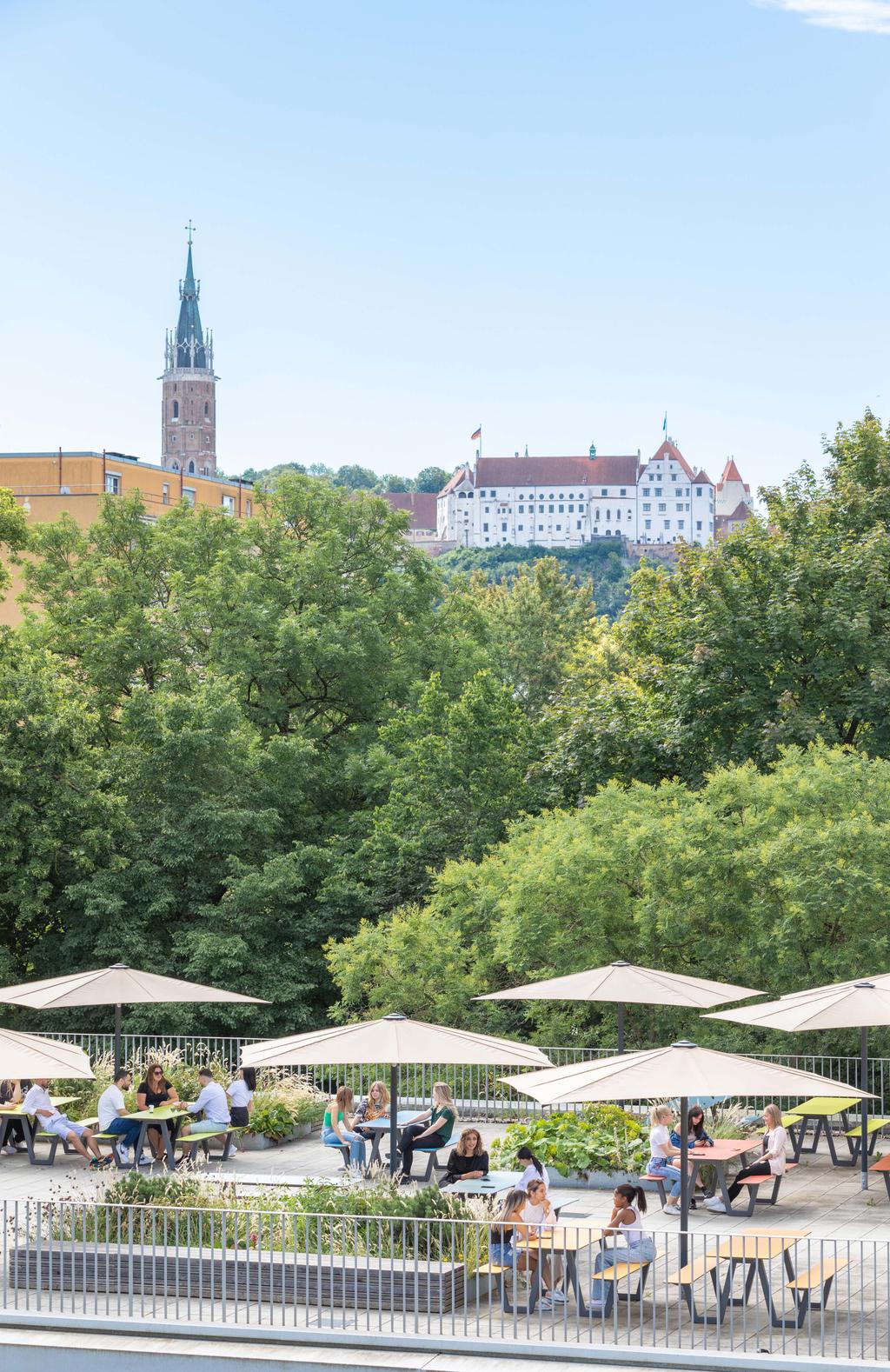 State vocational school, Landshuth with beige SCHATTELLO parasols A terrace with beige parasols where people are sitting, in the background a historic town with a church tower and castle