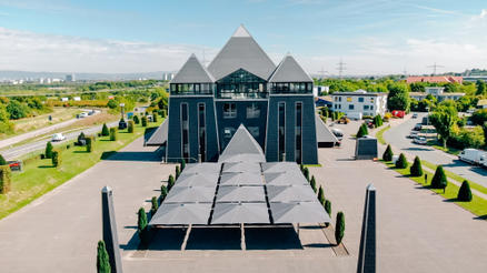 L’image montre un ensemble de bâtiments modernes avec une grande pyramide et d’autres constructions, devant lesquels plusieurs parasols gris sont alignés.