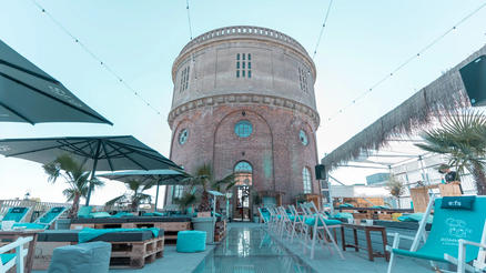 Salon d'été Dalwigk avec teintes bleu turquoise ALBATROS Une terrasse avec des chaises longues et des tables sous des parasols devant une tour ronde en briques.