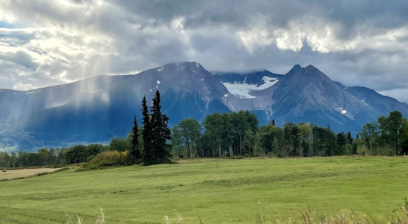 Hudson Bay Mountain with glacier in background, green field in foreground and stormy clouds with sun rays