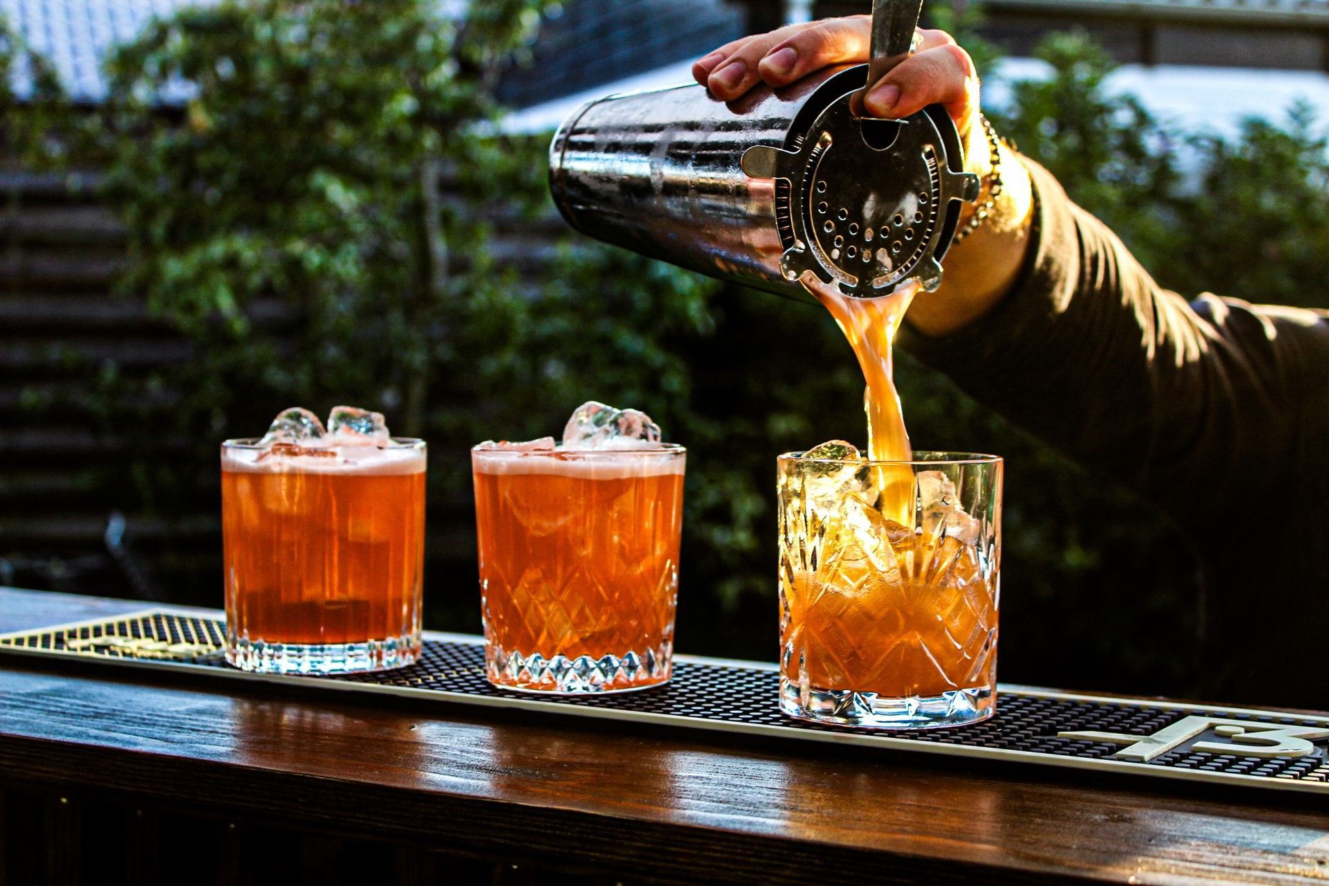 Bartender pours an orange cocktail from a shaker into a crystal glass with ice at an outdoor bar