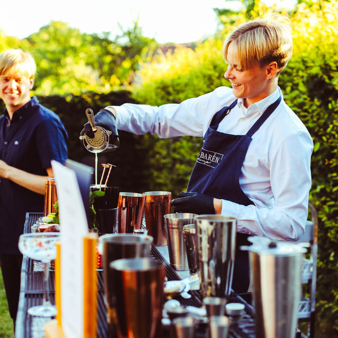 Bartender in a dark blue apron mixing drinks at an outdoor bar with cocktail shakers in sunshine