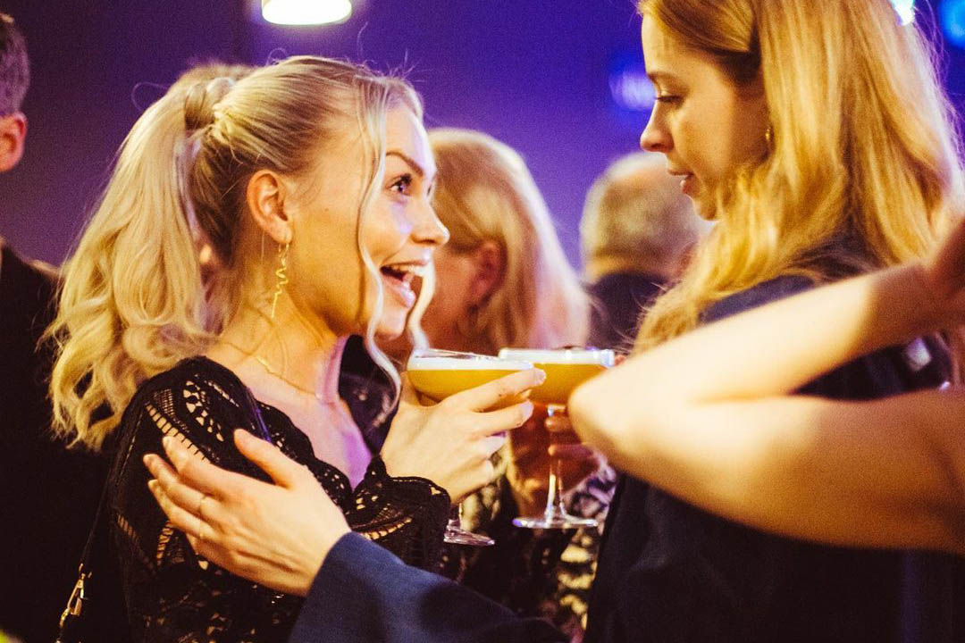 Two women cheerfully clinking cocktail glasses at an evening party with purple ambient lighting and guests in the background
