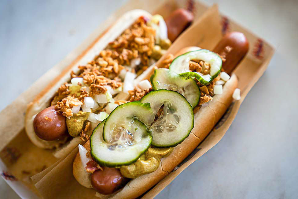 To hotdogs med sprøde løg, agurkeskiver, sennep og hakket løg i papbakke