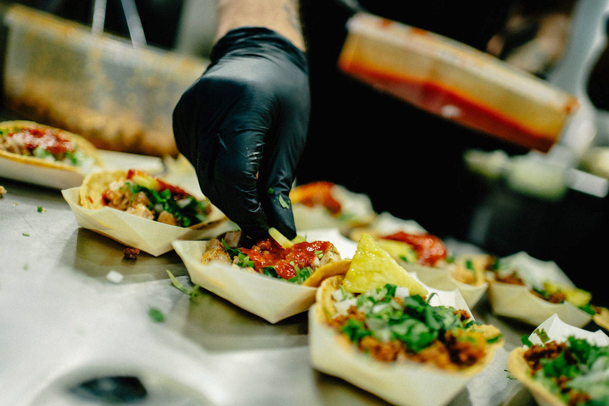 Chef in black glove tops tacos with salsa and coriander in paper trays