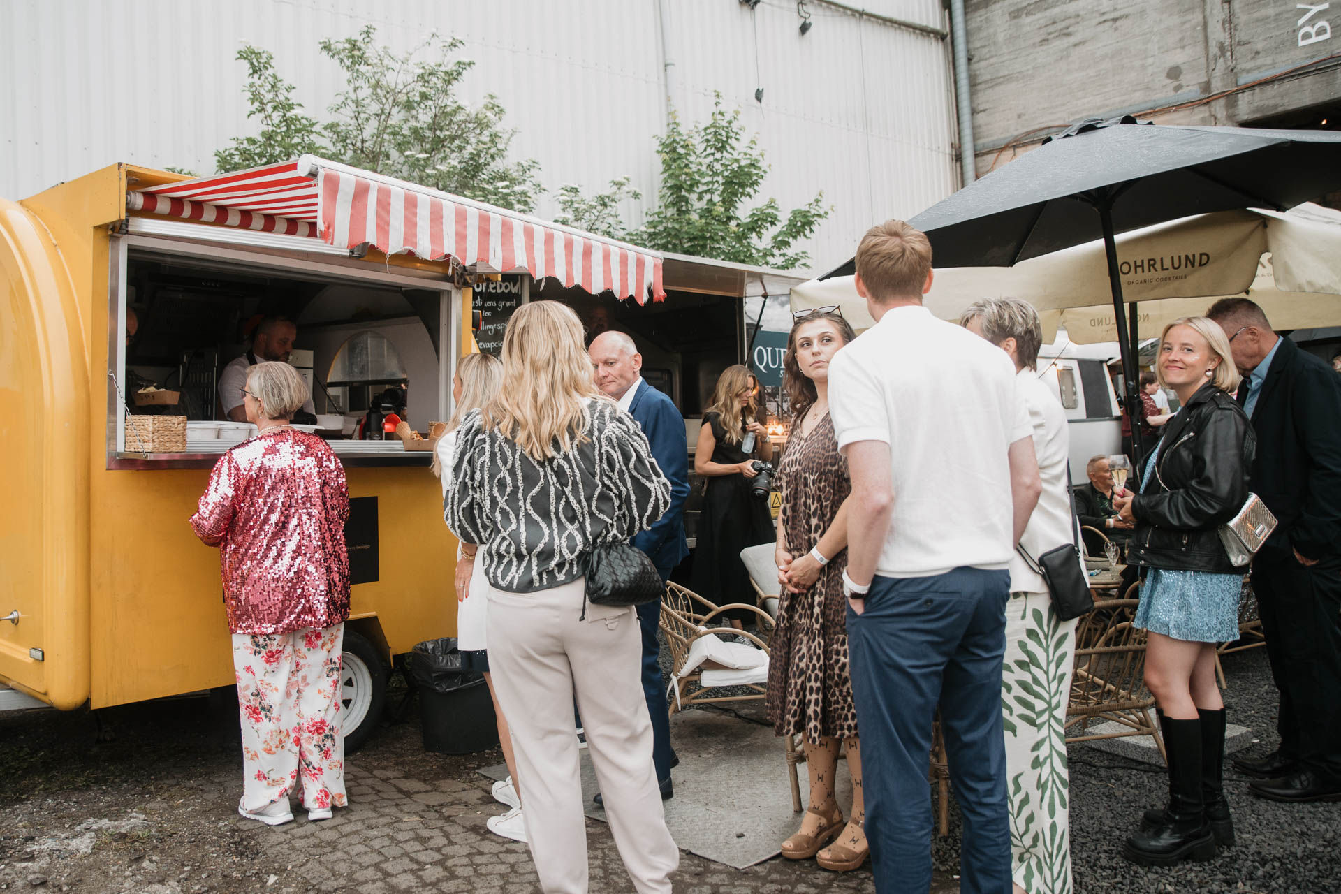 Gul foodtrailer med rød-hvid markise ved udendørs fest, mens gæster i festtøj står i kø og venter på mad