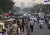 A busy urban street congested with traffic and street vendors. Numerous people walk along the roadside, while makeshift stalls selling fruits and other goods occupy the footpath. Vehicles, including cars, bikes, and auto-rickshaws, struggle to navigate through the crowd. Police officers are present, monitoring the situation. The background features tall buildings and streetlights, indicating a growing cityscape.