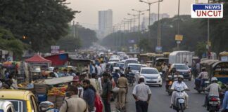 A busy urban street congested with traffic and street vendors. Numerous people walk along the roadside, while makeshift stalls selling fruits and other goods occupy the footpath. Vehicles, including cars, bikes, and auto-rickshaws, struggle to navigate through the crowd. Police officers are present, monitoring the situation. The background features tall buildings and streetlights, indicating a growing cityscape.