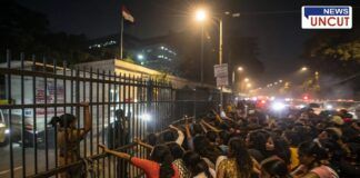 A large crowd of young women gathers outside a gated police recruitment center in Pune at night. Some are seen pushing against the iron gate while security personnel try to manage the situation. The dimly lit scene is illuminated by streetlights and vehicle headlights, with a tense atmosphere as the crowd struggles to enter. A woman in a red dress reaches out toward the gate, while others look on anxiously. The background features a government building with an Indian flag and traffic congestion, adding to the chaotic environment.