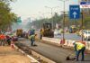 Road widening work underway in Pune to improve traffic flow. Workers and construction machinery are engaged in road development, with traffic and city buildings visible in the background. A directional sign indicating Pune is also seen in the image.
