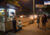 A nighttime street scene near Innovative School shows a small roadside stall selling tobacco products. A young schoolboy in uniform is purchasing an item from the vendor, while another boy sits nearby smoking. In the background, vehicles pass by on a busy road, and pedestrians, including a woman and a young girl, walk along the sidewalk. The dim lighting and informal setup highlight concerns about underage tobacco sales and public safety in the area.
