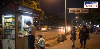 A nighttime street scene near Innovative School shows a small roadside stall selling tobacco products. A young schoolboy in uniform is purchasing an item from the vendor, while another boy sits nearby smoking. In the background, vehicles pass by on a busy road, and pedestrians, including a woman and a young girl, walk along the sidewalk. The dim lighting and informal setup highlight concerns about underage tobacco sales and public safety in the area.