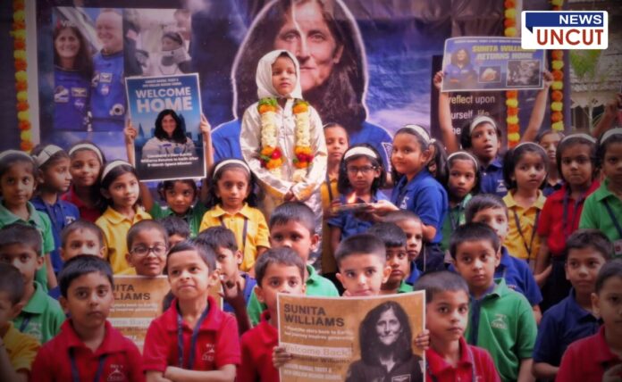 A group of Indian school children in colorful uniforms gathers to celebrate astronaut Sunita Williams' return to Earth. A young girl dressed as an astronaut, adorned with a flower garland, stands at the center, symbolizing Williams. Students hold posters and banners with Williams' images and messages like 