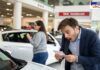 A shocked man in a blue blazer looking at a price receipt inside a car showroom. Behind him, a woman is examining a white car. A red sign in the background reads 'TAX INCREASE,' indicating rising vehicle costs.