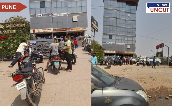 Outside view of Chakan Wines in Pune during Holi celebrations. People with colored faces are gathered near the entrance, some on motorcycles, while others stand in groups. The surroundings include a modern commercial building with a glass façade, parked vehicles, and a signboard of a nearby hotel. The scene captures the festive yet busy atmosphere of the area.