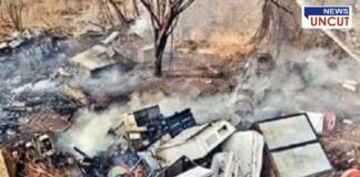 A scrapyard with burnt electronic waste and industrial debris, emitting smoke after a recent fire. Damaged refrigerators, metal scraps, and broken appliances are scattered across dry land. A charred tree stands in the background, and the ground is covered in ash. The "News Uncut" logo is visible in the top right corner.