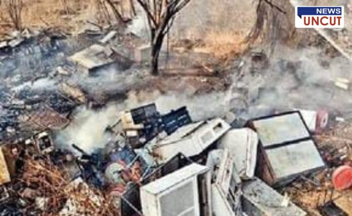 A scrapyard with burnt electronic waste and industrial debris, emitting smoke after a recent fire. Damaged refrigerators, metal scraps, and broken appliances are scattered across dry land. A charred tree stands in the background, and the ground is covered in ash. The 