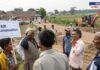 Villagers protesting against land acquisition for the Hinjewadi-Man road widening project. A young protester holds a sign demanding 'Fair Compensation,' while local residents argue with an official at the construction site. An excavator and partially built road are visible in the background, highlighting the ongoing development.