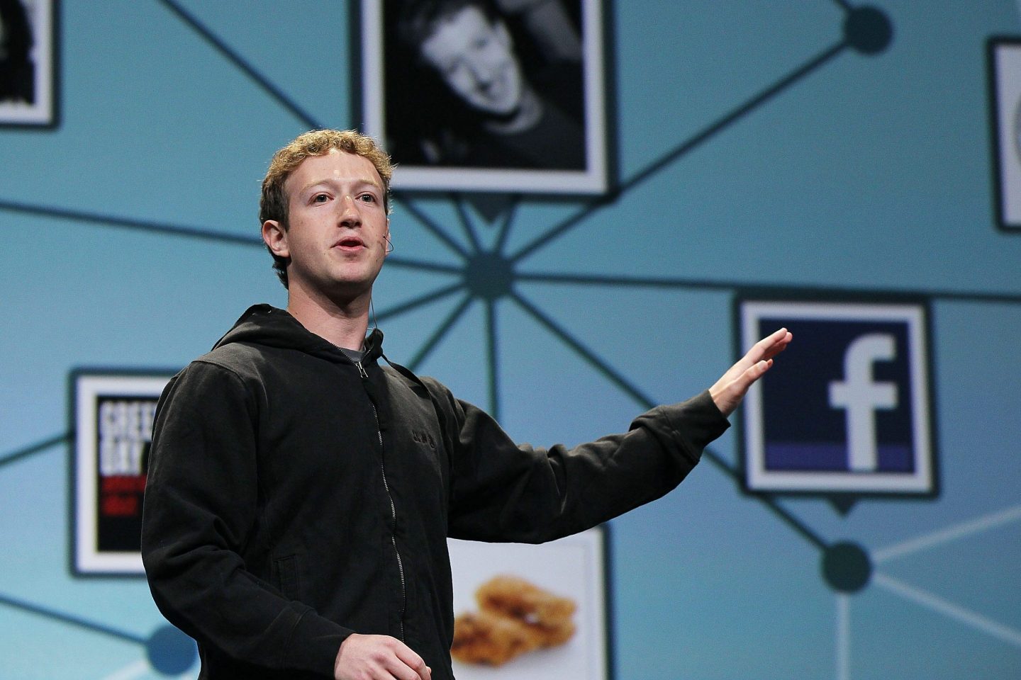 A man in a black zip-up stands gesturing out to his left in front of background with a Facebook logo