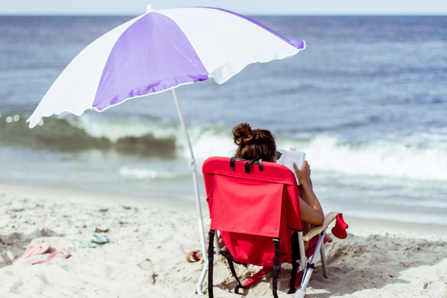 A girl reading on the beach and sitting on a red beach chair with a parasol