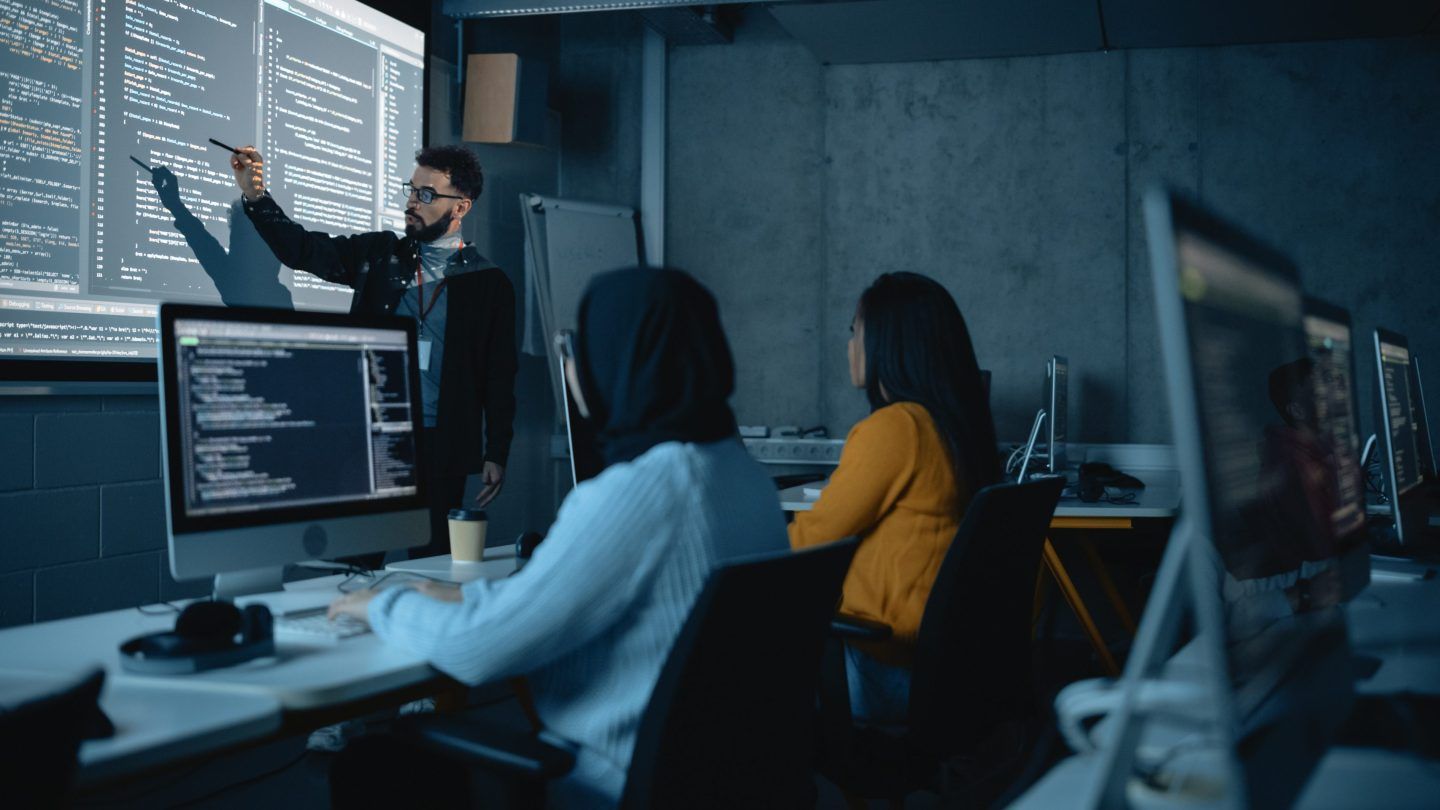 Teacher at the front of a classroom teaches coding, which is displayed behind him on a projector. Two students sit at a table watching.