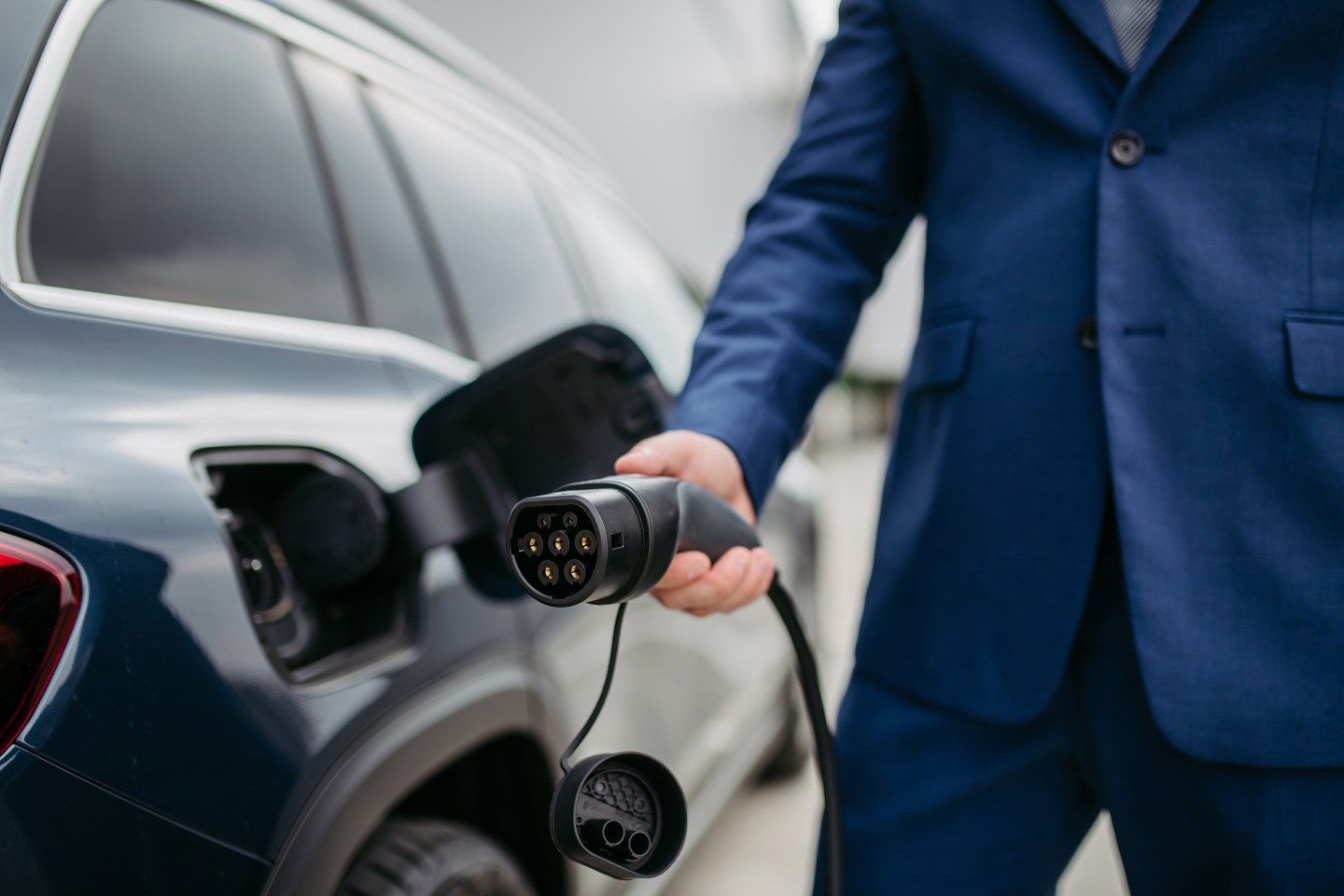 A close-up of businessman charging his electric car at charging station in front of his office. Detail of man's hand plugging in power cord to an electric vehicle.