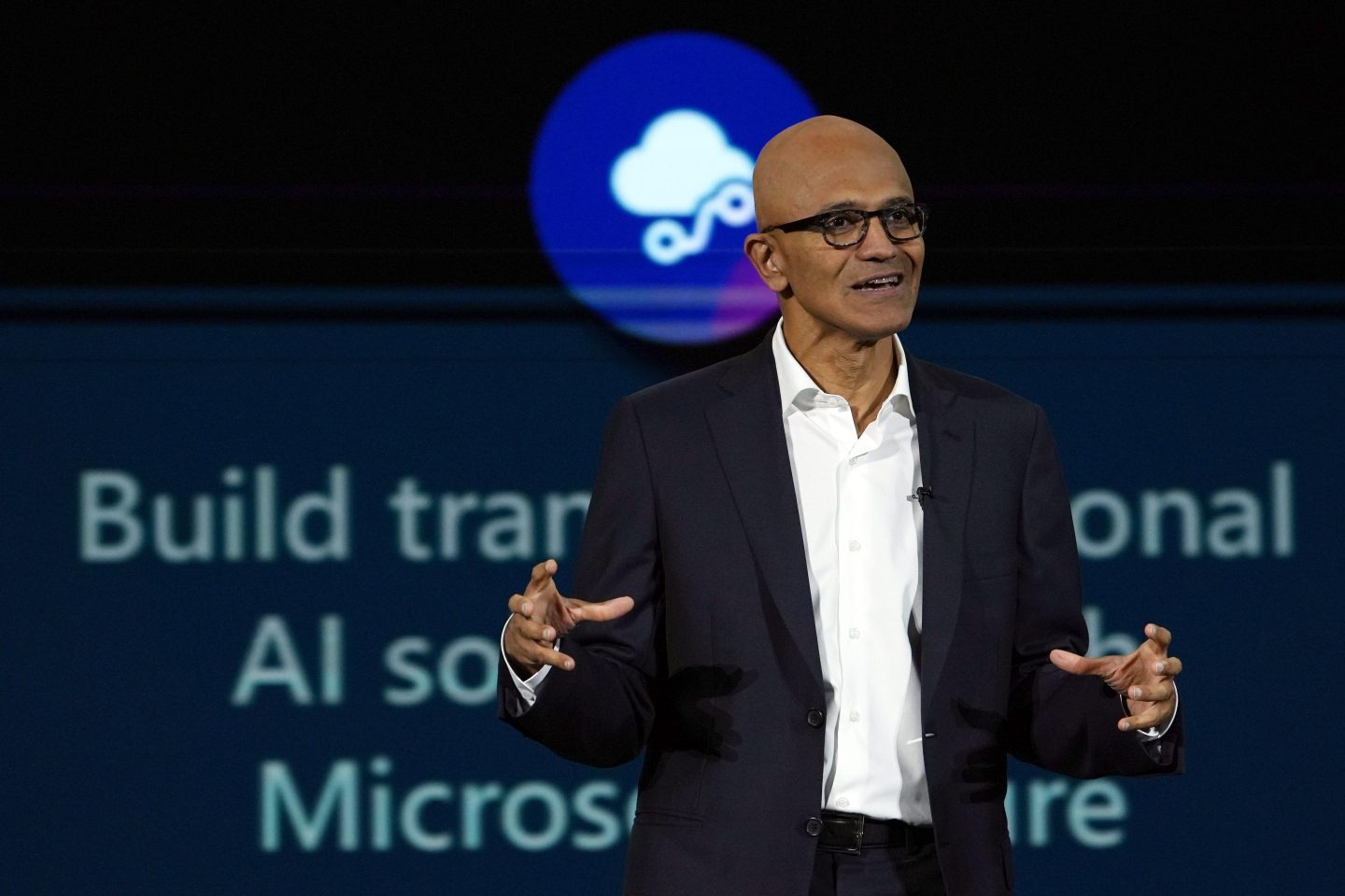 Satya Nadella smiles and lifts his arms by his side as he speaks on stage.