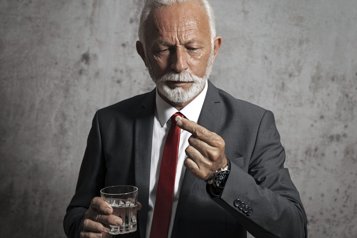 An older man wearing a business suit holds a pill in one hand and a glass of water in the other.