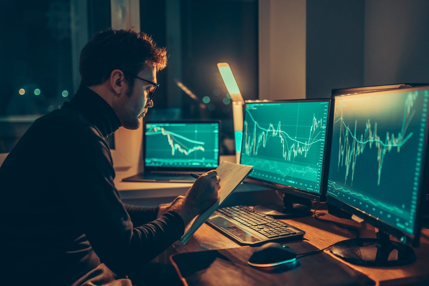 Man looks at market trends in front of three monitors at night.