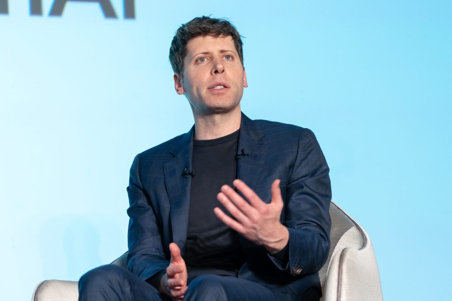 OpenAI CEO Sam Altman on stage sitting in a chair gesturing with his left hand.