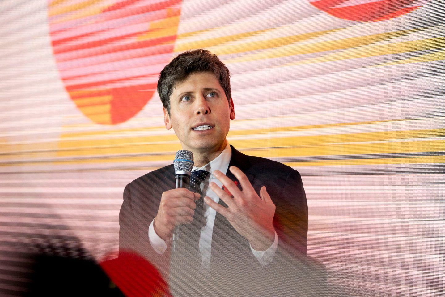Sam Altman holds a microphone and speaks amid a bright multicolor backdrop.