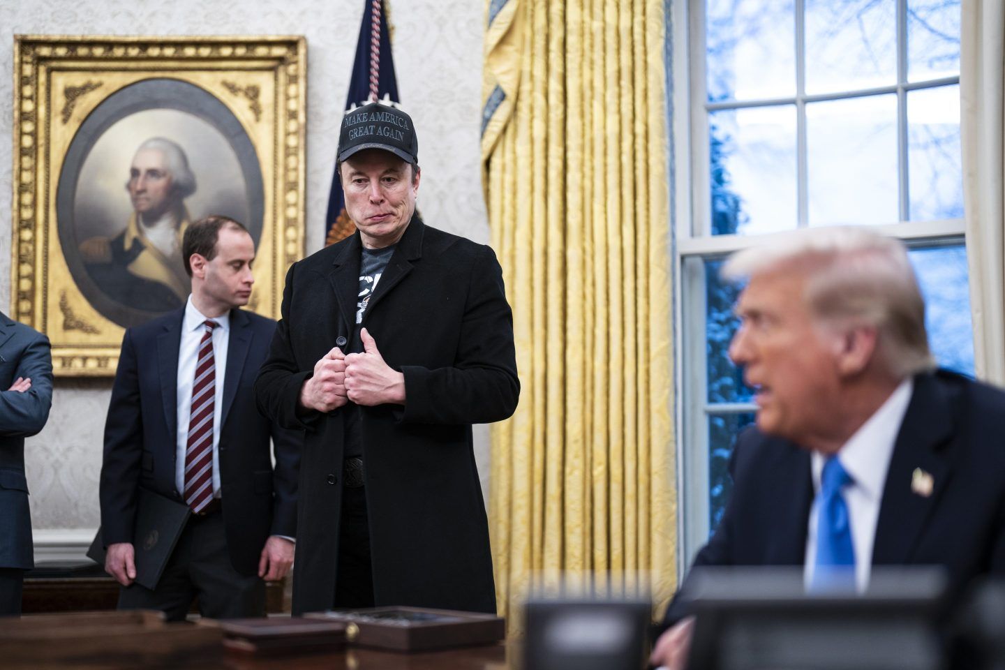 Elon Musk stands in the Oval Office and looks over at Donald Trump, who is sitting behind his desk.
