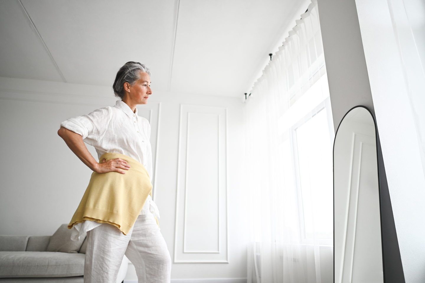 Middle aged woman dressed in white looking into a full-length mirror