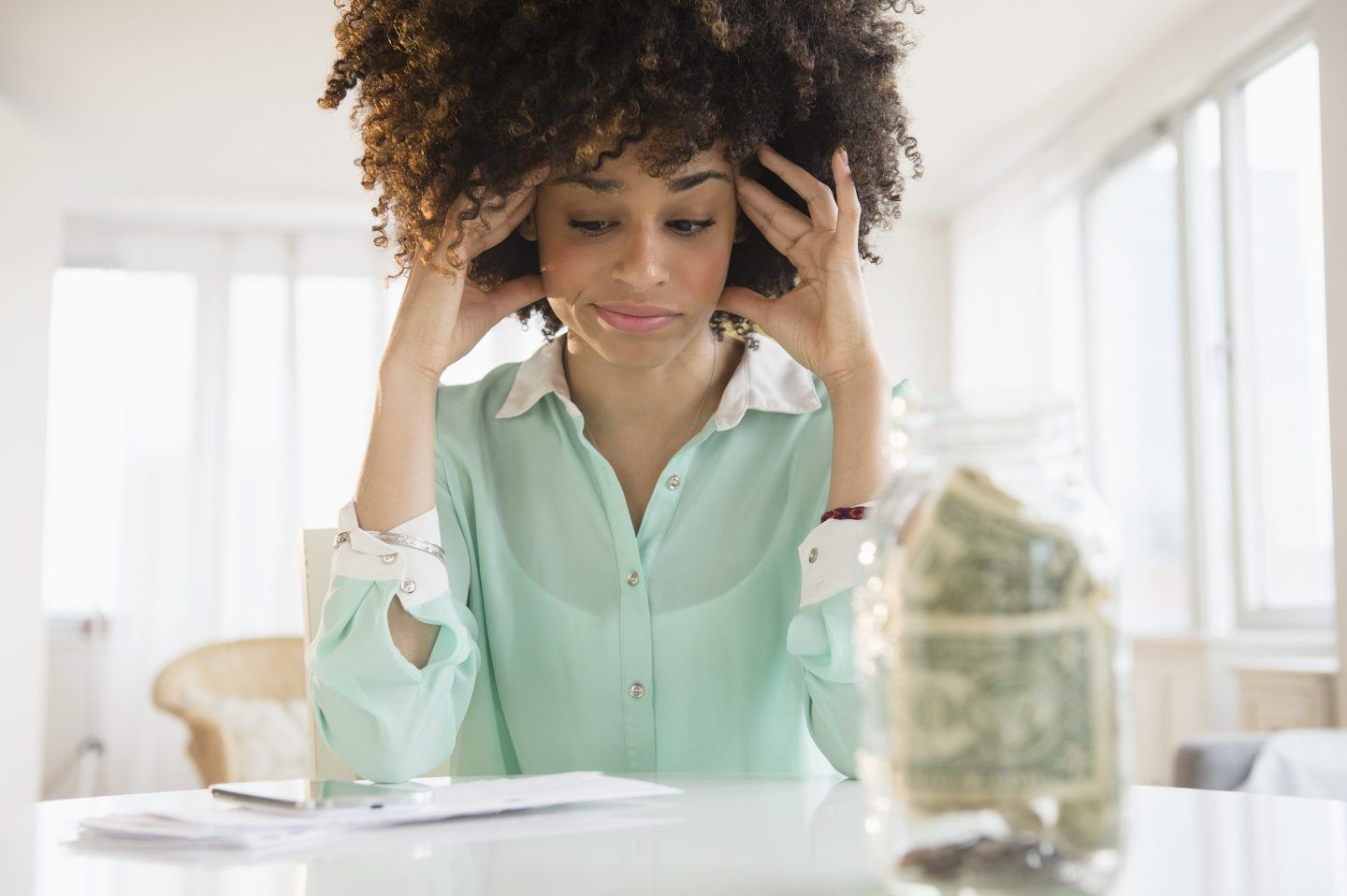 A young woman stares at a pile of bills in front of her, looking concerned. A mason jar of cash sits in the foreground.