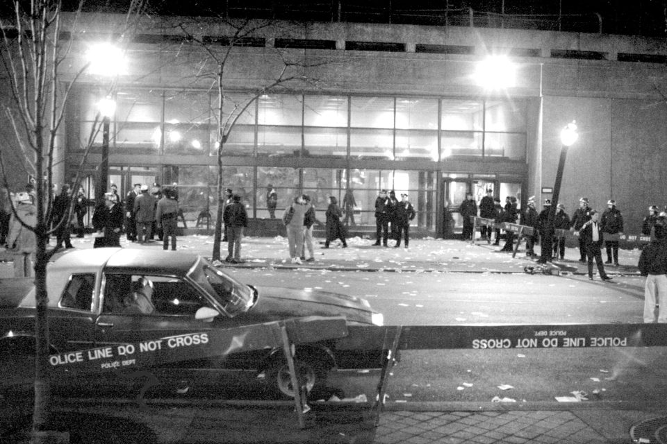 The outside of a gymnasium building has people standing around outside and is blocked off with police barricades.