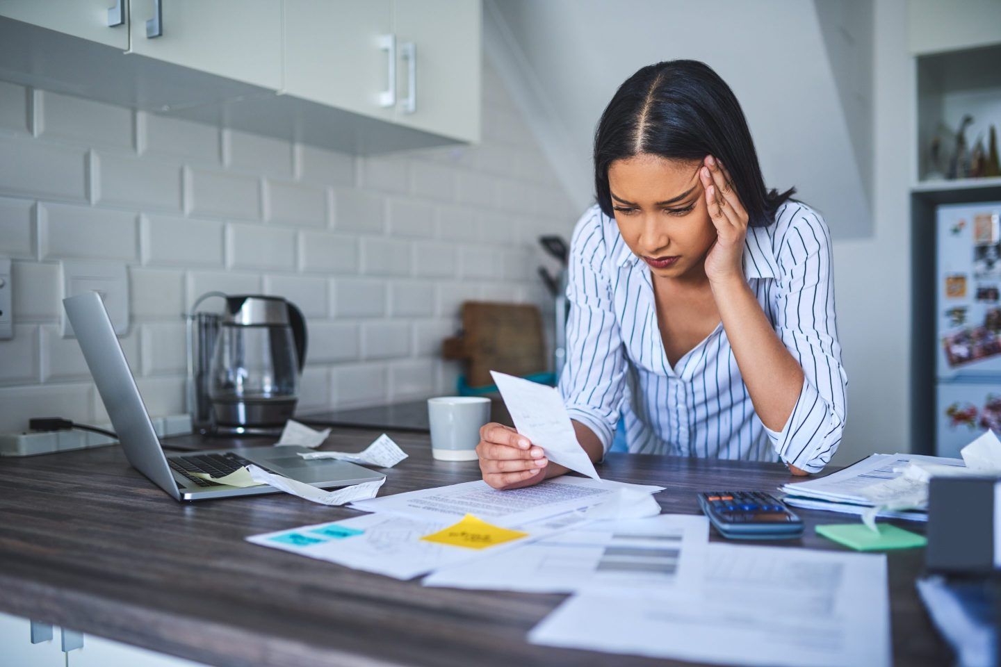 Young businesswoman stands in a kitchen looking stressed while looking at scattered papers