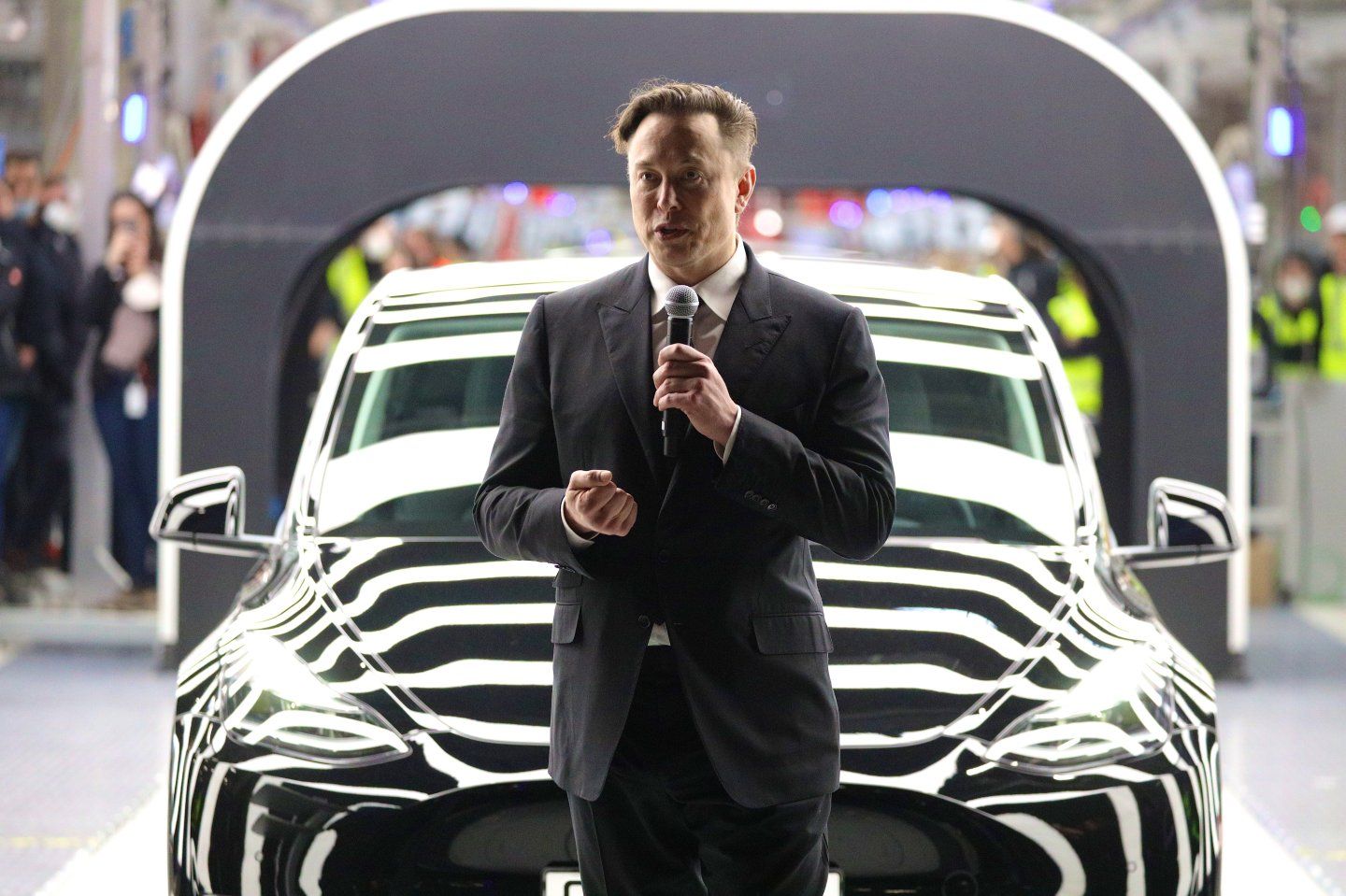 Elon Musk speaks into a microphone in front of a Tesla EV.