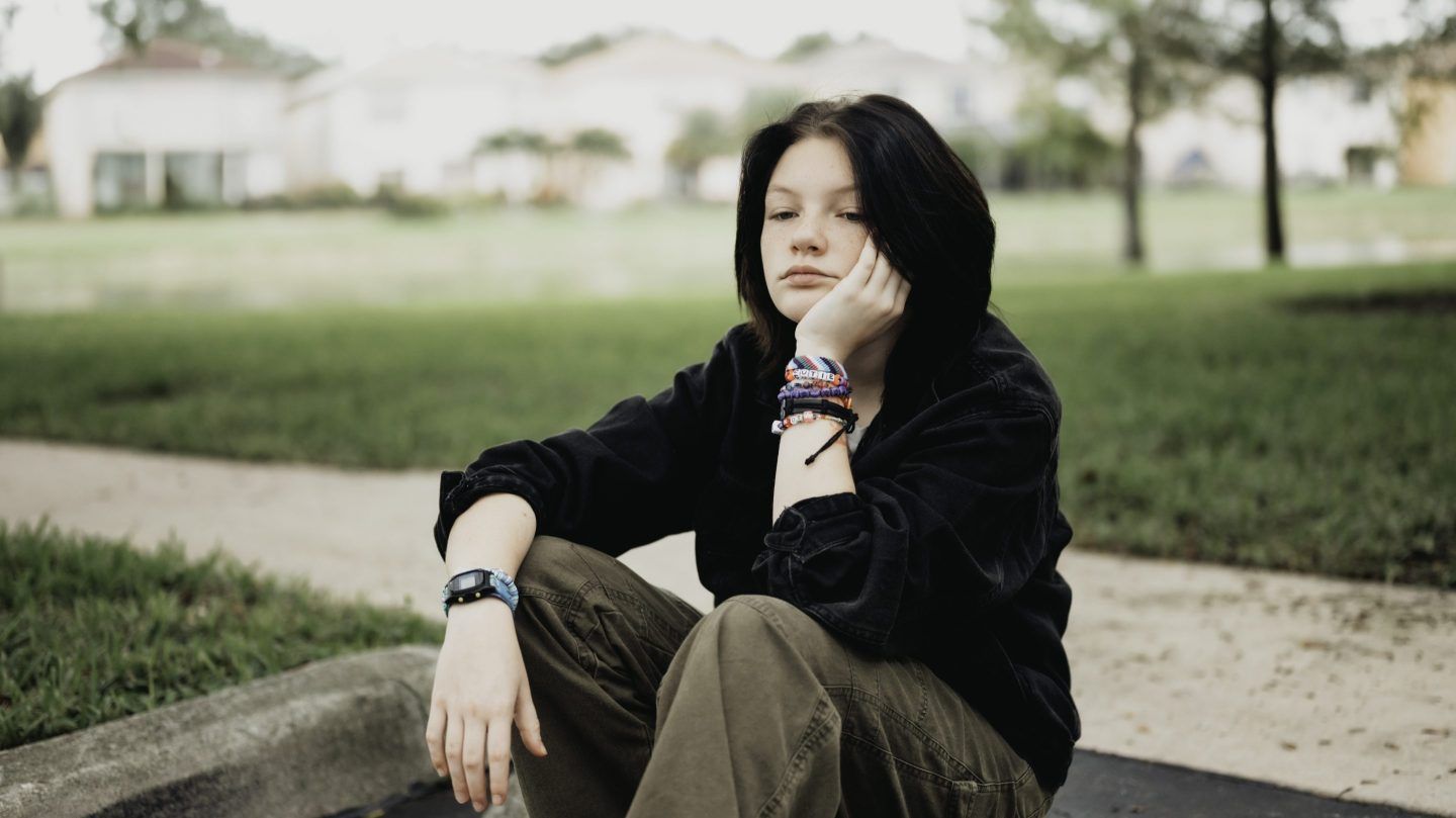 Sad teen in black sweatshirt and woven bracelets sitting on a curb