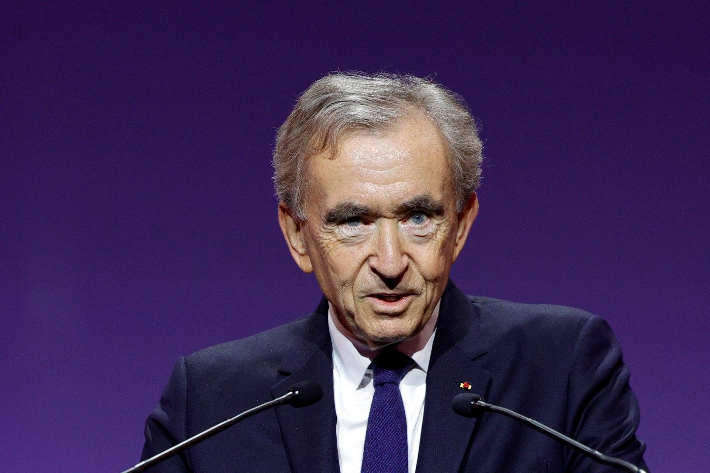 Bernard Arnault, wearing a dark suit, speaks into a microphone and is in front of a deep purple background.