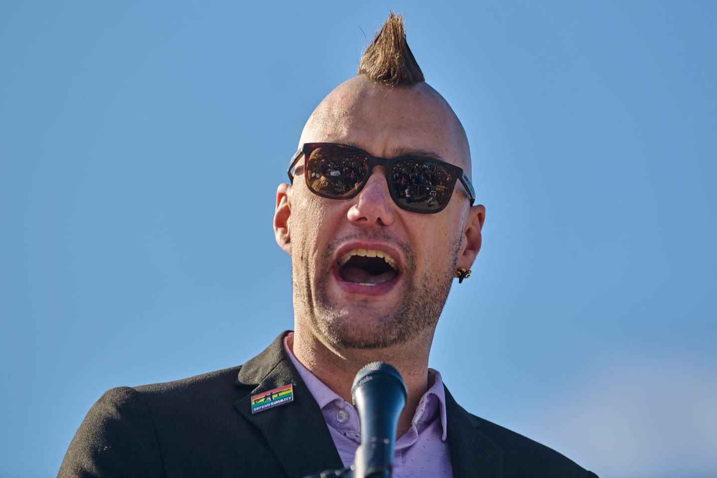 Nate Brough, with a mohawk and sunglasses, speaks outside with a blue sky behind him.