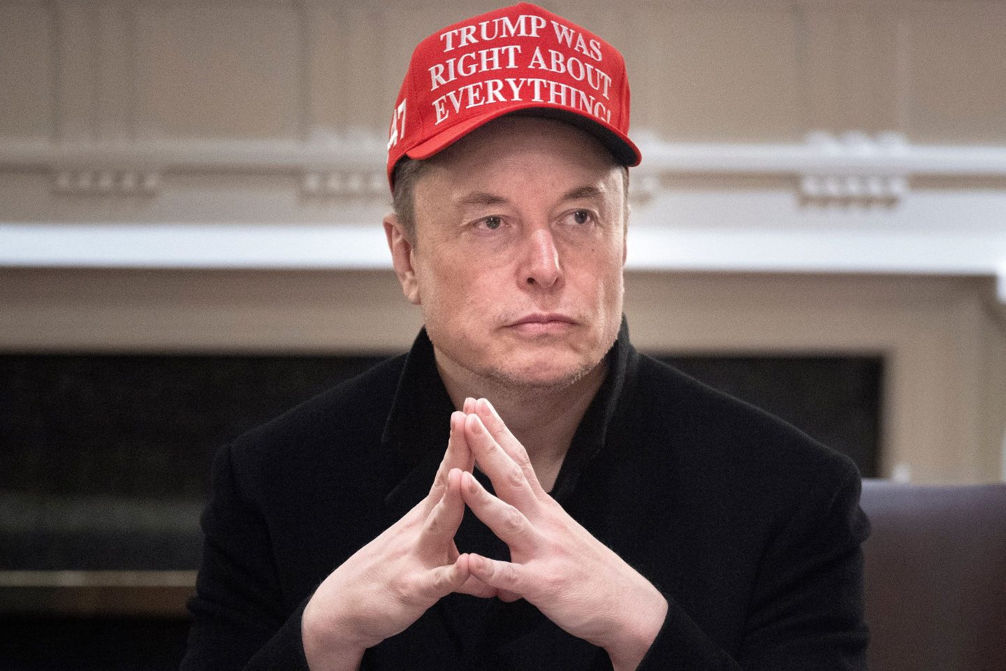Elon Musk presses his hands together on a table. He is wearing a red "Trump was right about everything" hat.