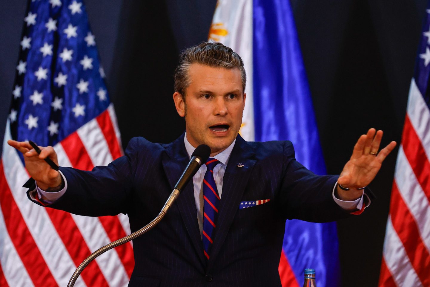 Pete Hegseth, standing in front of a line of American flags, speaks and gestures broadly with both hands.