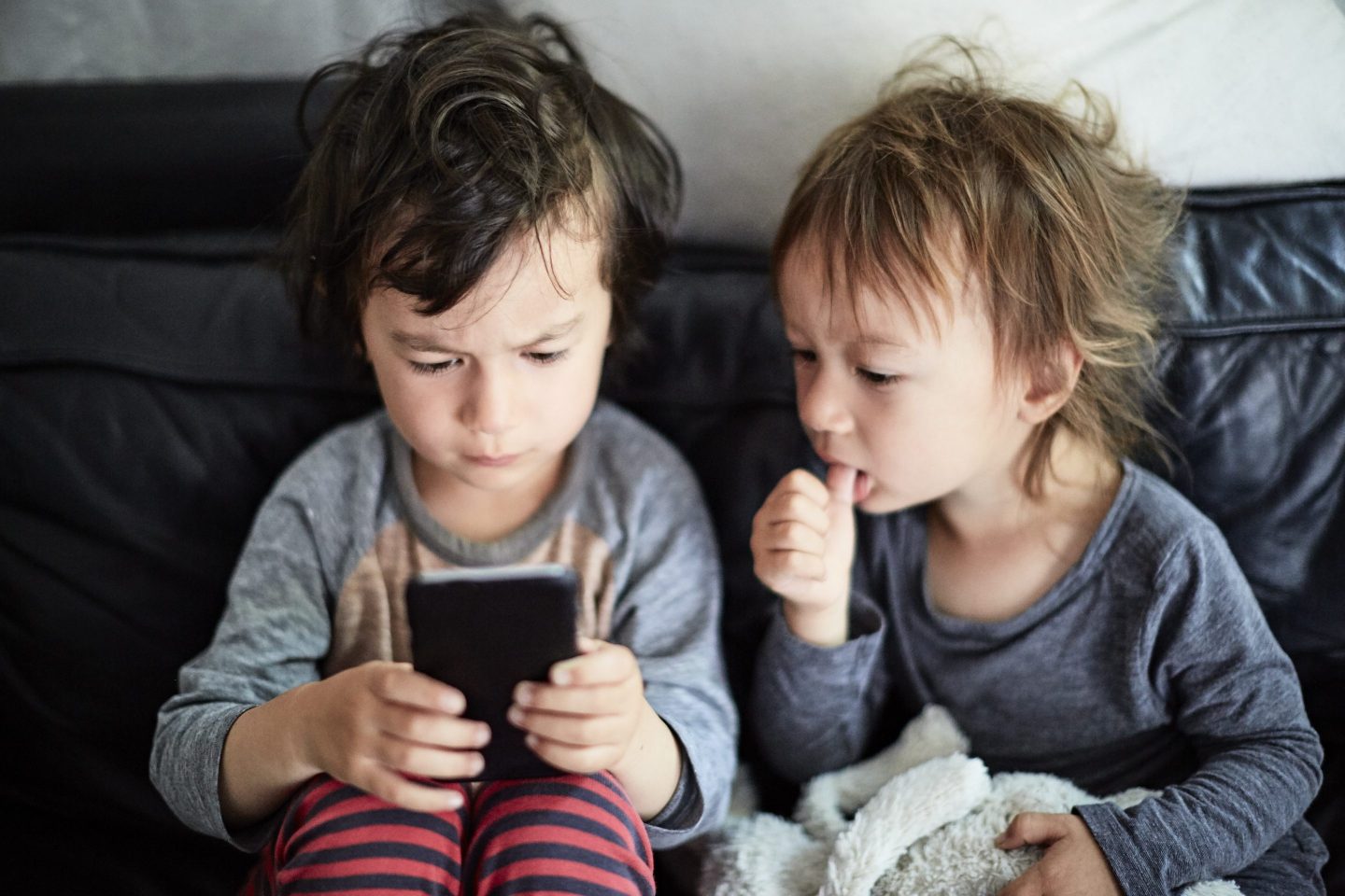 Two tiny boys look at a smartphone, as one sucks his thumb