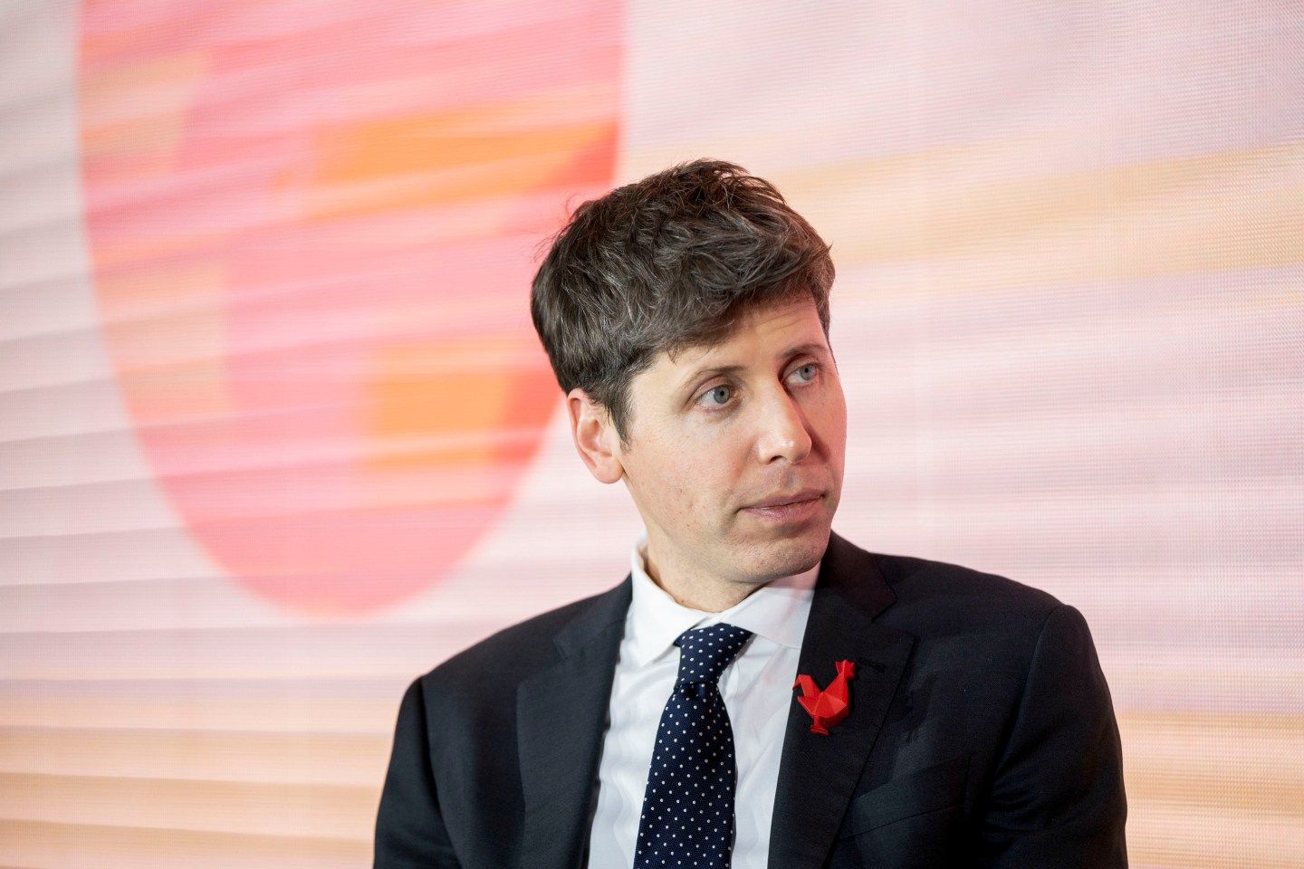 Sam Altman on a stage in a suit in front of a pink background.