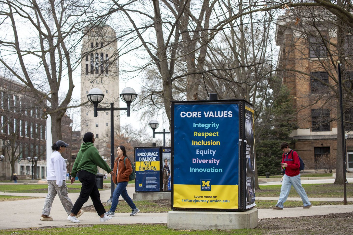 University of Michigan students walk on the UM campus next to signage displaying the University's "Core Values"