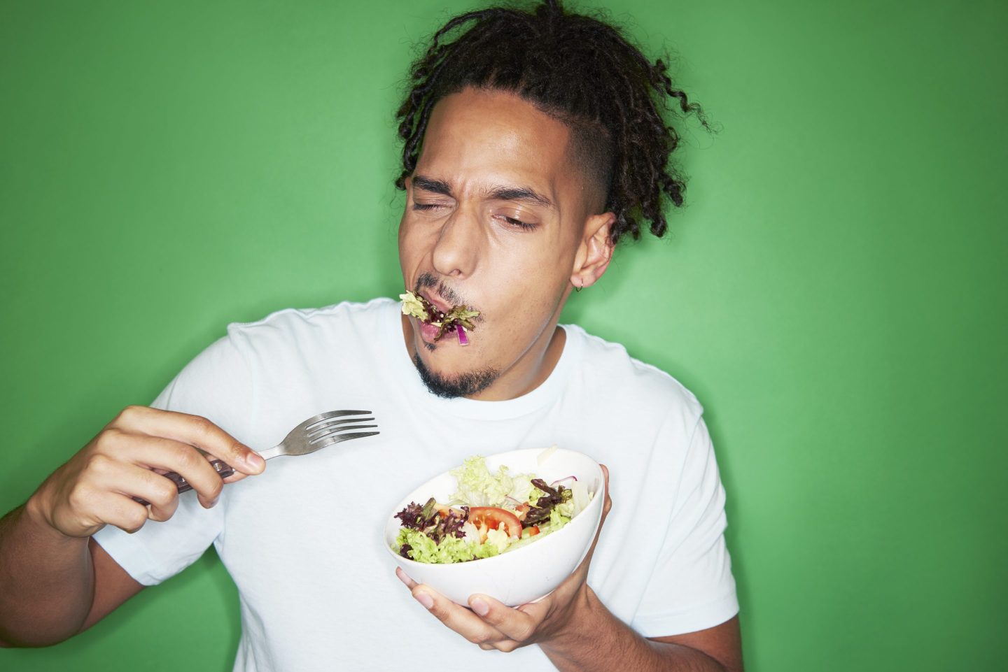 Man in white T-shirt holding a white bowl of salad in one hand and a fork in the other, chewing some salad