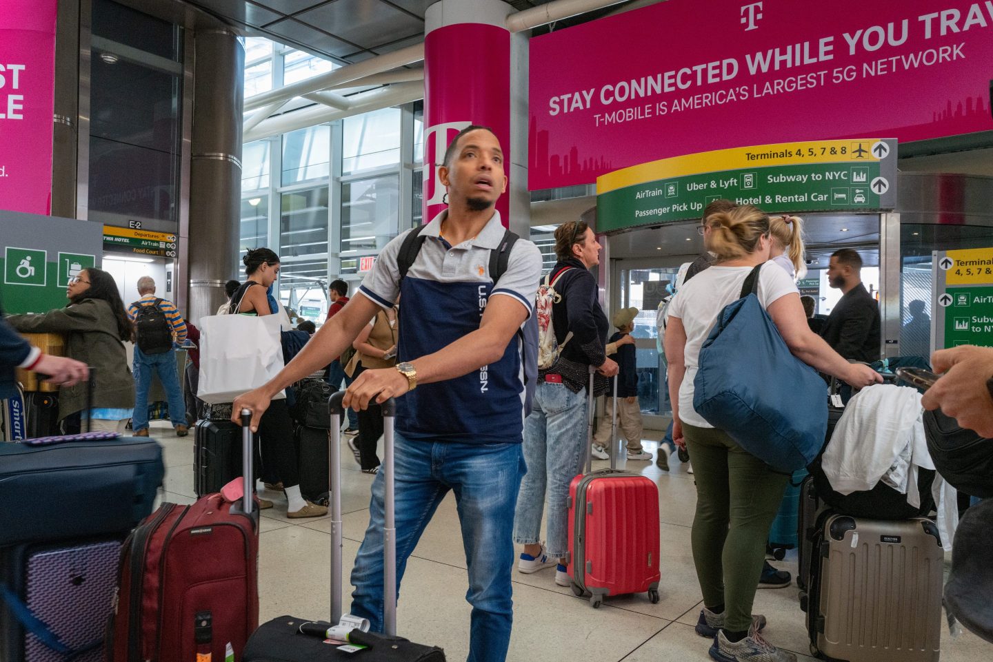 A man holding the handles of two suitcases looks intently at something as people move through a crowded JFK International Airport.